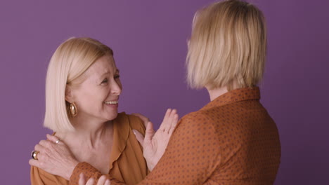 Two-Blonde-Mature-Women-Wearing-Brown-Shirt-Hugging-And-Smiling-On-Purple-Background