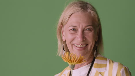 Blonde-Mature-Woman-With-Blue-Eyes-Dressed-In-Striped-Shirt-And-Accessories-Posing-Smelling-A-Sunflower-And-Smiling-At-Camera-On-Green-Background