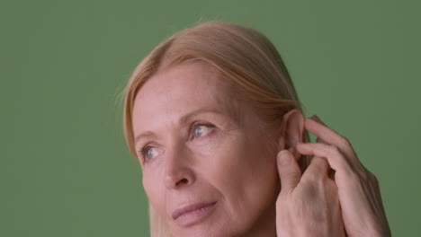 Close-Up-View-Of-Blonde-Mature-Woman-Wearing-Green-Shirt,-Picking-Her-Hair-Behind-Her-Ear-Looking-At-Side-On-Green-Background