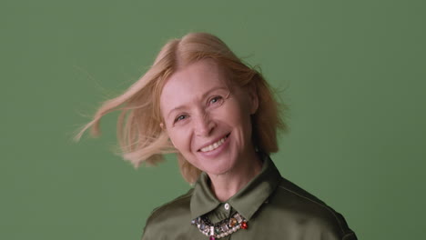 Blonde-Reife-Frau-Mit-Grünem-Hemd-Lächelt-In-Die-Kamera,-Während-Der-Wind-Ihre-Haare-Und-Ihr-Hemd-Auf-Grünem-Hintergrund-Bewegt