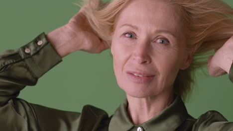 Nahaufnahme-Einer-Blonden-Reifen-Frau-Mit-Grünem-Hemd,-Die-In-Die-Kamera-Lächelt-Und-Ihr-Haar-Mit-Wind-Auf-Grünem-Hintergrund-Berührt