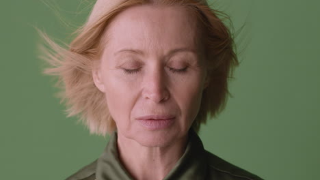 Close-Up-View-Of-Blonde-Mature-Woman-Wearing-Green-Shirt-With-Closed-Eyes-While-The-Wind-Move-Her-Hair-On-Green-Background,-Then-She-Looks-At-Camera