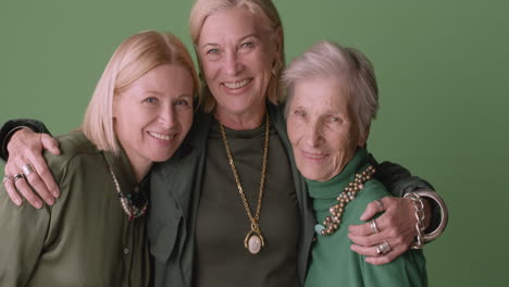 Two-Blonde-Mature-Women-And-Blonde-Senior-Woman-Smiling-And-Hugging,-Wearing-Green-Tones-Clothes-And-Posing-On-Green-Background