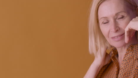 Fröhliche-Blonde-Reife-Frau-Mit-Braunem-Hemd-Tanzt,-Lächelt-Und-Berührt-Ihr-Haar-Auf-Orangefarbenem-Und-Violettem-Hintergrund
