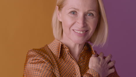 Nahaufnahme-Einer-Blonden-Reifen-Frau-Mit-Braunem-Hemd,-Die-Lächelt-Und-Ihre-Schulter-Auf-Orangefarbenem-Und-Violettem-Hintergrund-Berührt