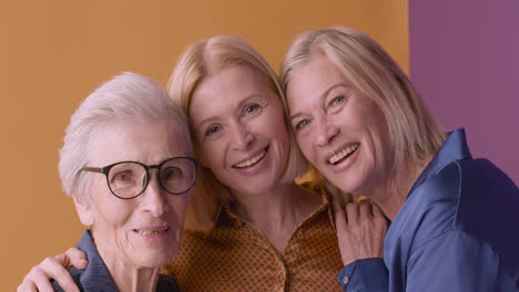Two-Blonde-Mature-Women-And-Blonde-Senior-Woman-Smiling-And-Hugging-Posing-On-Orange-And-Purple-Background