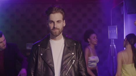 Portrait-Of-Handsome-Bearded-Man-In-Leather-Jacket-Smiling-At-Camera-At-Disco-While-His-Friends-Dancing-Behind-Him