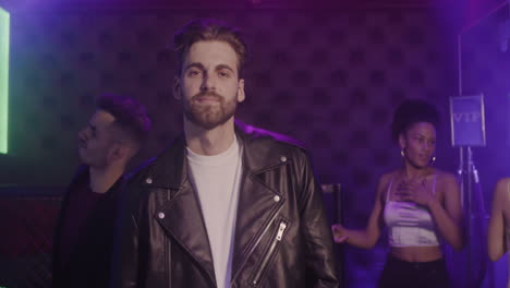 Portrait-Of-Handsome-Bearded-Man-In-Leather-Jacket-Smiling-At-Camera-At-Disco-While-His-Friends-Dancing-Behind-Him-1