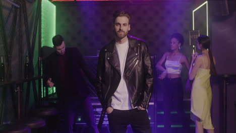 Handsome-Bearded-Man-In-Leather-Jacket-Smiling-At-Camera-At-Disco-While-His-Friends-Dancing-Behind-Him