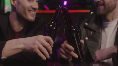 Close-Up-Of-Two-Male-Friends-Toasting-With-Beer-Bottle-And-Talking-Together-While-Sitting-At-Bar-Counter