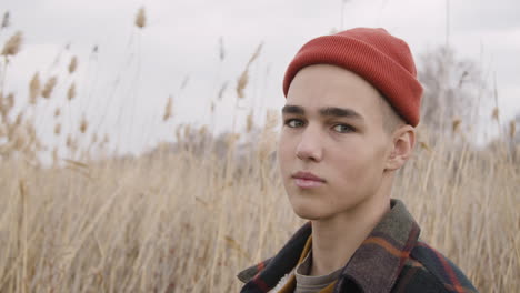 Close-Up-View-Of-A-Teenage-Boy-With-Orange-Beanie-And-Plaid-Coat-Looking-At-Side-In-A-Wheat-Field,-Then-Looks-At-Camera-And-Smiles