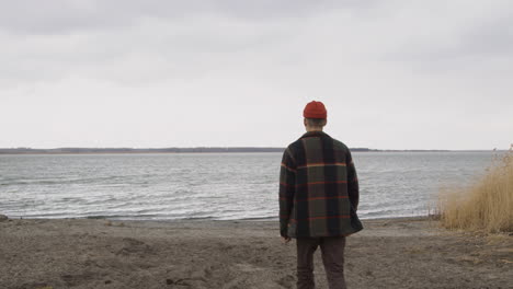 Rear-View-Of-A-Teenage-Boy-Dressed-In-An-Orange-Beanie-And-A-Plaid-Coat-Approaching-The-Seashore