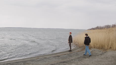 Teenage-Boy-Dresses-In-An-Orange-Beanie-And-Plaid-Coat-Standing-Near-Of-Seashore-On-A-Cloudy-Day,-Then-A-Teenage-Girl-In-Winter-Clothes-Approachs-Him