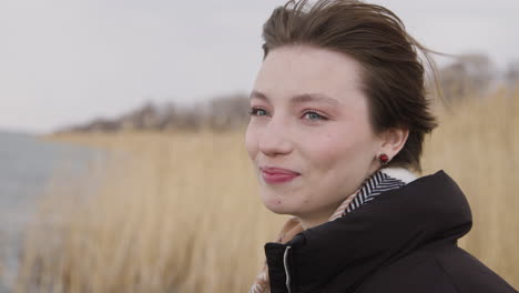 Close-Up-View-Of-Teenage-Girl-With-Short-Hair-Wearing-Scarf-And-Coat-Looking-At-Camera-And-Smiling-On-A-Windy-Day