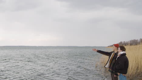 Teenage-Boy-And-Teenage-Girl-Standing-Near-Of-Seashore-On-A-Cloudy-Day,-Boy-Show-Something-With-His-Arm
