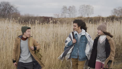 Two-Teenage-Boy-And-Girl-And-Teenager-Boy-With-Long-Hair-Wearing-Winter-Clothes-Walking-In-A-Wheat-Field-On-A-Cloudy-Day