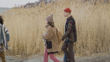 Side-View-Of-A-Teenage-Boy-And-Girl-Approaching-To-Two-Friends-Wearing-Winter-Clothes-Walking-In-A-Wheat-Field-Near-Of-Sea-On-A-Cloudy-Day