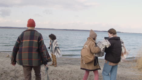 Group-Of-Teenage-Boys-And-Girls-Wearing-Winter-Clothes-Walking-Towards-The-Seashore-With-Backpacks-While-Taking-Photos-Of-Their-Friends-On-A-Cloudy-Day