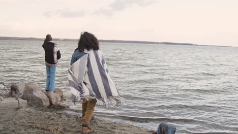 Rear-View-Of-Two-Friends-In-Winter-Clothes-Throwing-Pebbles-To-The-Water-On-A-Seashore-On-A-Cloudy-Day