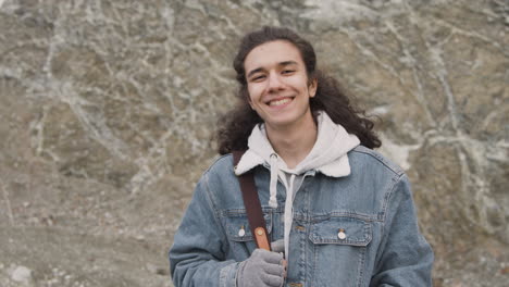 Front-View-Of-A-Teenage-Boy-With-Long-Hair-And-Winter-Clothes-Smiling-And-Looking-At-Camera-On-The-Mountain-On-A-Windy-Day