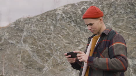 Teenage-Boy-With-Orange-Beanie-And-Winter-Clothes-Looking-At-Smartphone-On-The-Mountain
