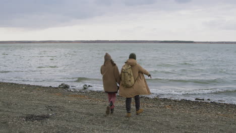 Rear-View-Of-Two-Friends-In-Winter-Clothes-Walking-To-The-Seashore-On-A-Windy-Day