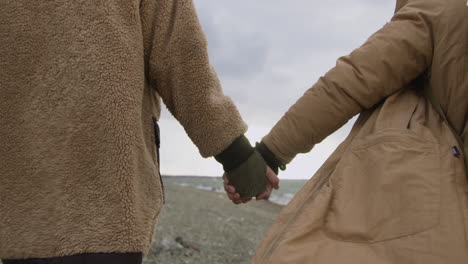 Rear-View-Of-Two-Teenage-Holding-Hands-In-Winter-Clothes-Walking-On-Seashore