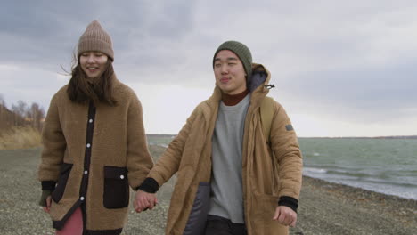 Front-View-Of-Two-Teenage-Holding-Hands-In-Winter-Clothes-Talking-And-Walking-On-Seashore