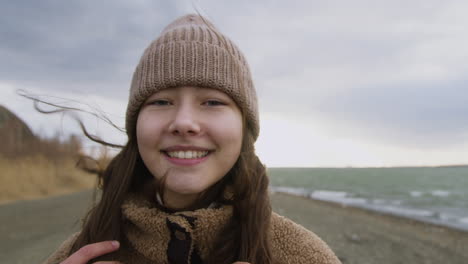 Close-Up-View-Of-Teenage-Girl-Looking-At-Camera-And-Smiling-On-Seashore-On-Windy-Day