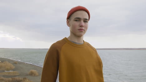 Front-View-Of-A-Teenage-Boy-With-Orange-Beanie-And-Sweatshirt-Posing-At-Camera-On-The-Top-Of-The-Mountain-On-The-Beach-On-A-Windy-Day