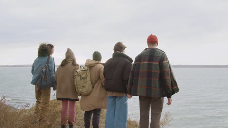 Rear-View-Of-Group-Of-Teenage-Friends-In-Winter-Clothes-Holding-Hands-And-Raising-Arms-On-Top-Of-The-Mountain-On-The-Beach-On-A-Windy-Day