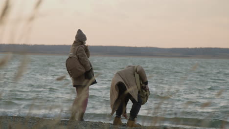 Rear-View-Of-Two-Friends-In-Winter-Clothes-Throwing-Pebbles-To-The-Water-On-A-Seashore-On-A-Cloudy-Day-1