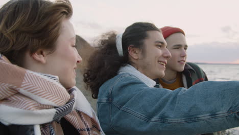 Close-Up-View-Of-Three-Teenage-Friends-In-Winter-Clothes-On-Seashore-Talking-And-Looking-The-Ocean-On-A-Windy-Day
