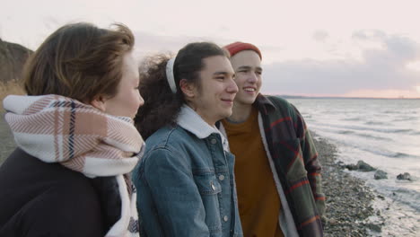 Drei-Jugendliche-Freunde-In-Winterkleidung-An-Der-Küste-Reden,-Lächeln-Und-Schauen-An-Einem-Windigen-Tag-Auf-Den-Ozean