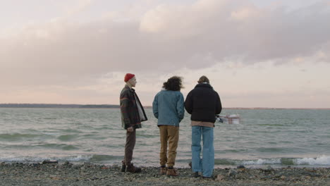 Rear-View-Of-Three-Friends-In-Winter-Clothes-Talking-And-Throwing-Pebbles-To-The-Water-On-A-Seashore-On-A-Cloudy-Day