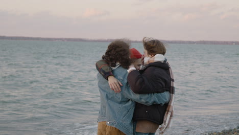Three-Friends-In-Winter-Clothes-Hugging-And-Smiling-On-A-Seashore-On-A-Windy-Day