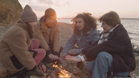 Gruppe-Jugendlicher-Freunde,-Die-Sich-Am-Lagerfeuer-Neben-Der-Küste-Unterhalten-Und-Sich-Die-Hände-Wärmen-1
