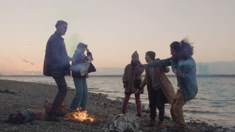 Gruppe-Von-Teenager-Freunden,-Die-Um-Ein-Lagerfeuer-Tanzen,-Das-Bunte-Wunderkerzen-An-Der-Küste-Hält