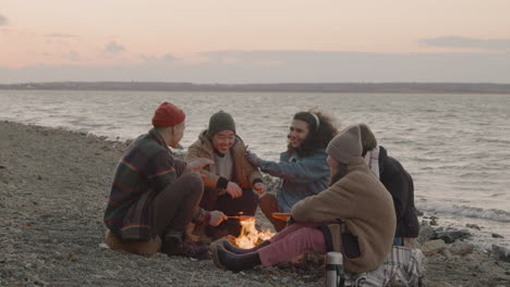 Group-Of-Teenage-Friends-Sitting-Around-The-Bonfire-While-They-Roasting-Sausages-And-Talking-On-The-Seashore