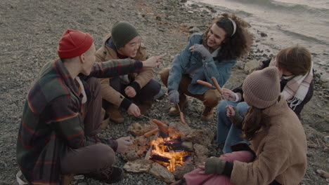 Top-View-Of-Group-Of-Teenage-Friends-Sitting-Around-The-Bonfire-While-They-Roasting-Sausages-And-Talk