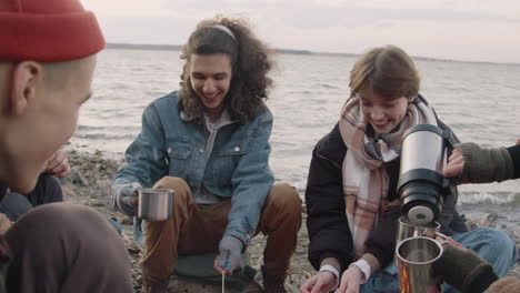 Group-Of-Teenage-Friends-Sitting-Around-The-Bonfire-While-They-Serving-Tea-In-Cups-With-A-Thermos-On-The-Seashore