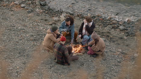 Group-Of-Teenage-Friends-Sitting-Around-The-Bonfire-While-They-Serving-Tea-In-Cups-With-A-Thermos-On-The-Seashore-1