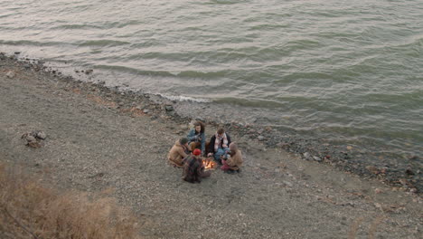 Top-View-Of-Group-Of-Teenage-Friends-Sitting-Around-The-Bonfire-While-Drinking-Tea-And-Roasting-Sausages-On-The-Seashore