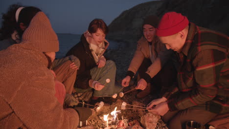 Eine-Gruppe-Jugendlicher-Freunde,-Die-Am-Lagerfeuer-Sitzen,-Während-Sie-Marshmallows-Rösten-Und-Sich-Am-Meeresufer-Unterhalten