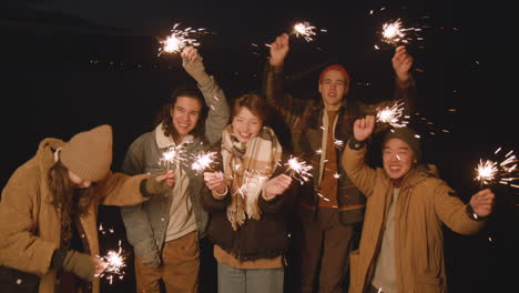 Close-Up-View-Of-Group-Of-Friends-Dressed-In-Winter-Clothes-Waving-At-The-Camera-Holding-Sparklers-At-Night