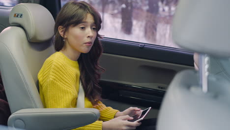 Camera-Focuses-On-Girl-Sitting-On-The-Back-Seat-Using-A-Tablet-While-Talking-To-Their-Parents