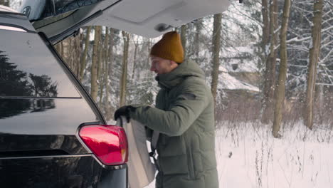 Side-View-Of-A-Man-Dressed-In-Winter-Clothes-Putting-Things-Into-The-Trunk-Of-A-Car-In-A-Snowy-Forest