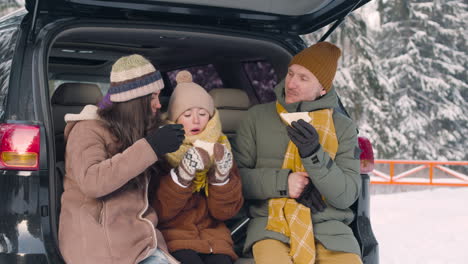 Parents-And-Daughter-Eating-Sandwiches-And-Drinking-Hot-Drink-Sitting-In-The-Trunk-Of-The-Car-In-A-Snowy-Forest