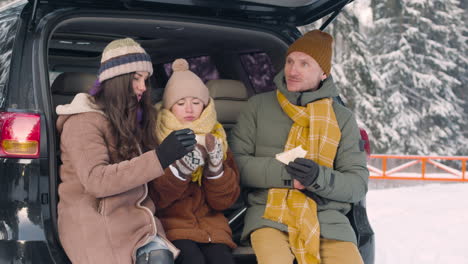 Parents-And-Daughter-Eating-Sandwiches-And-Drinking-Hot-Drink-Sitting-In-The-Trunk-Of-The-Car-In-A-Snowy-Forest-1
