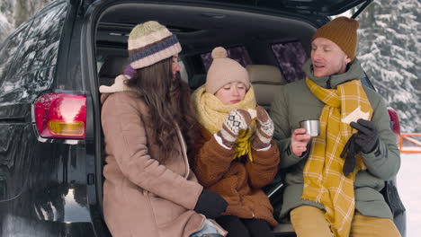 Parents-And-Daughter-Eating-Sandwiches-And-Drinking-Hot-Drink-Sitting-In-The-Trunk-Of-The-Car-In-A-Snowy-Forest-2